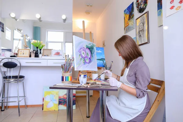 Dziewczyna siedzi na stołku sztalugi i piśmie malarstwa, używa pędzla do — Zdjęcie stockowe
