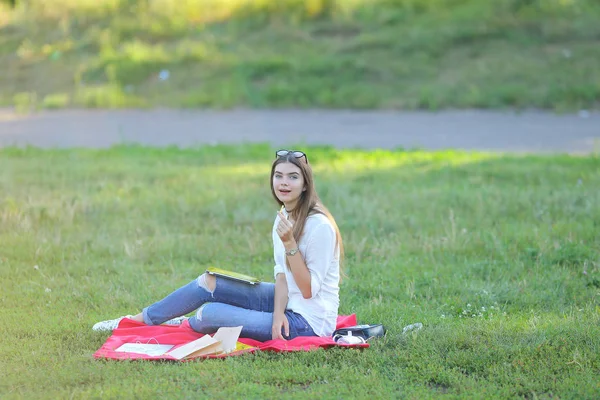 Giovane ragazza seduta sull'erba nel parco e lavora in un computer portatile e mangiare fast food — Foto Stock