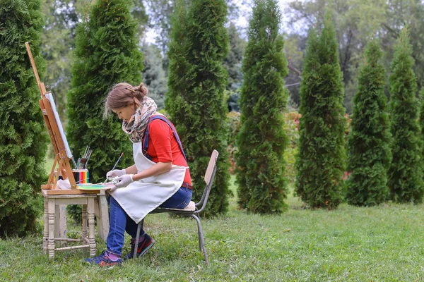 Κορίτσι καλλιτέχνης ζωγραφίζει την εικόνα και κάθεται στην καρέκλα στο καβαλέτο πλευρές του t — Φωτογραφία Αρχείου