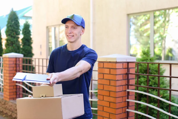 Cartero llevando un paquete en las manos sonriendo a la cámara en segundo plano — Foto de Stock