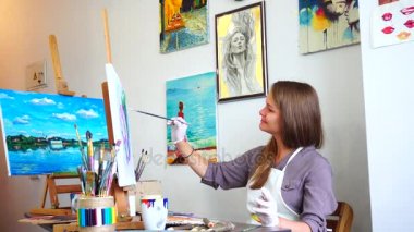 Kız sanatçı profilinde kamera için oturur ve resmi, bakarak darbeleri ve çizgiler şövale parlak sanat dersi Studio, fırça ile tuval üzerine çizer.