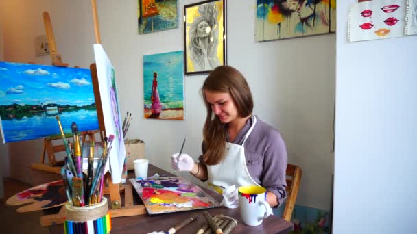 Κορίτσι καλλιτέχνης χρώματα σε καμβά. Ζωγράφος αναμιγνύει χρώμα στην παλέτα και βούρτσα απεικονίζει λουλούδια που κάθεται σε καρέκλα στο τραπέζι στην τάξη τέχνη. — Αρχείο Βίντεο