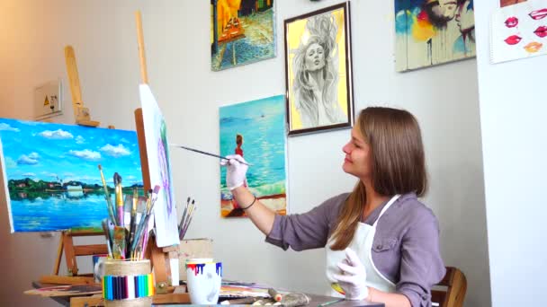 Девушка-художник сидит в профиле к камере и смотрит на картину, рисует мазки кисти и линии на холсте с помощью кисти на Easel в студии Яркий художественный класс . — стоковое видео