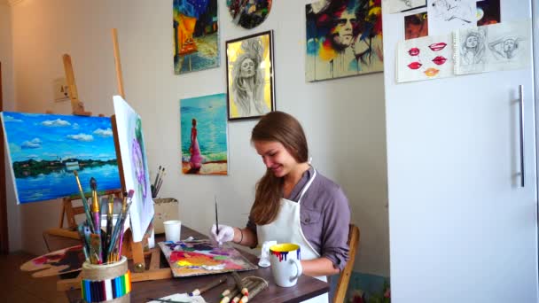 Künstlerin bringt Ende des Blumenmusters und posiert für die Kamera, während sie im Kunstatelier vor dem Hintergrund weißer Wände und farbiger Bilder sitzt. — Stockvideo