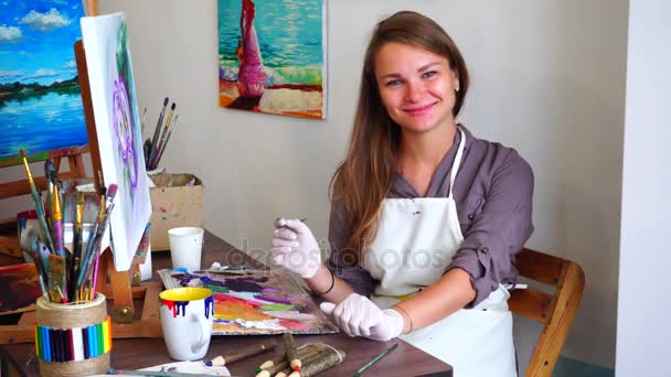 Artista bonita menina olha para a câmera e sorrindo enquanto segurando escova na mão e desenha imagem no cavalete sentado na cadeira no estúdio de arte . — Vídeo de Stock