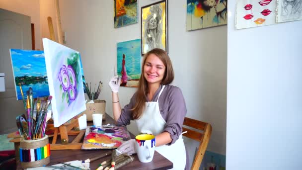 Porträt einer Künstlerin lächelt und lacht, blickt in die Linse eines Fotografen und sitzt an der Staffelei im Kunstatelier. — Stockvideo