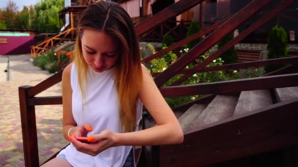 Jolie jeune fille tient le téléphone en main, échangé des messages, lit des SMS et des sourires, assis sur le banc sur fond de parterres de fleurs et d'escaliers à l'extérieur  . — Video