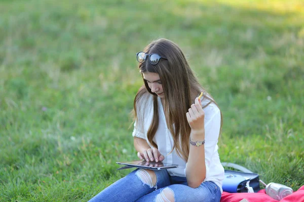 Niña sentada en la hierba en el parque y trabaja en un ordenador portátil y comer comida rápida — Foto de Stock