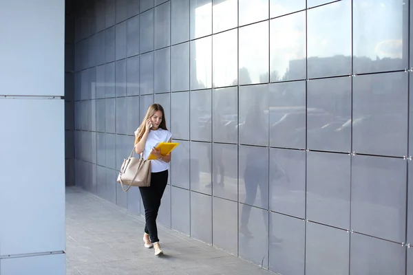Νεαρή γυναίκα περπατά και μιλά στο τηλέφωνο, κατέχει έγγραφα σε — Φωτογραφία Αρχείου