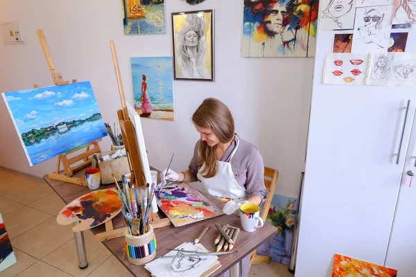 Dziewczyna siedzi na stołku sztalugi i piśmie malarstwa, używa pędzla do — Zdjęcie stockowe