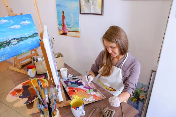 Chica se sienta en el taburete en el caballete y escribir pintura, utiliza el pincel para — Foto de Stock