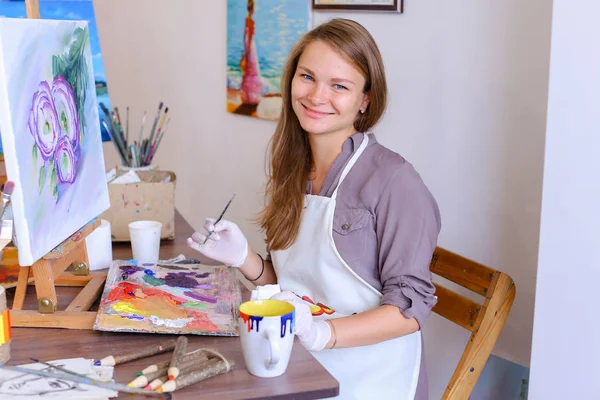 Κορίτσι που κάθεται σε σκαμνί στο καβαλέτο και ζωγραφική γράφοντας, χρησιμοποιεί τη βούρτσα για να — Φωτογραφία Αρχείου