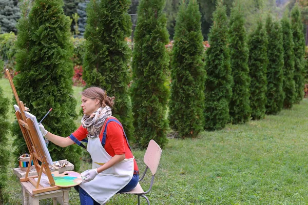 Ragazza artista dipinge quadro e si siede sulla sedia ai lati cavalletto di t — Foto Stock