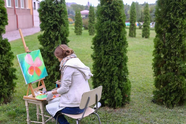 Dziewczyna artysta maluje obraz i siedzi na krześle po bokach sztalugi t — Zdjęcie stockowe