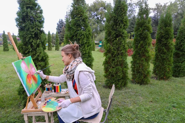 Menina artista pinta quadro e senta-se na cadeira em lados cavalete de t — Fotografia de Stock