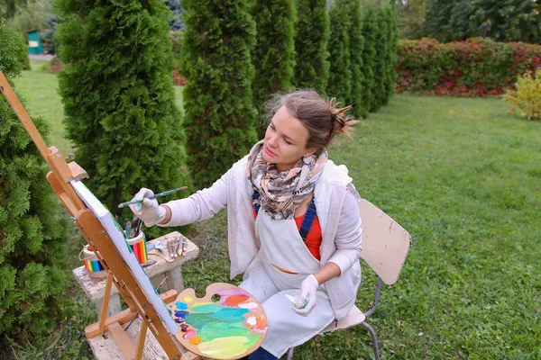 Chica artista pinta cuadro y se sienta en la silla a los lados del caballete de t —  Fotos de Stock