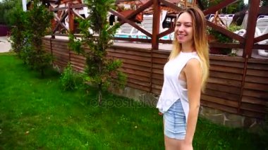 Hangi kamera önünde bakmak ve saç, düzeltiyor sevimli kız portresi gülümsüyor ve çit havuz ve açık havada yeşil bitki arka plan üzerinde yürüyor.