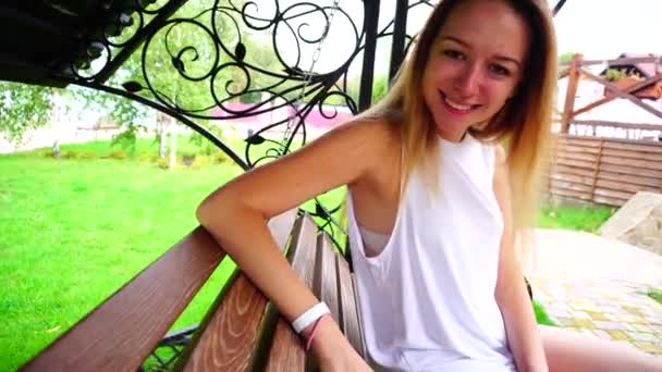 Retrato de la linda joven sentada y mecedora en el banco, mirando la cámara sonriendo y recreación al aire libre Día . — Vídeo de stock