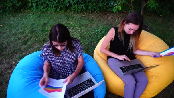Девушки начинающие предприниматели разрабатывают план работы на следующий месяц с помощью гаджета и сидя в открытом парке во второй половине дня . — стоковое видео