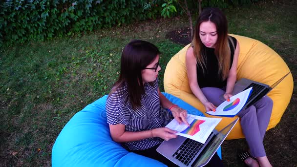 Des femmes financières mignonnes présentent des modifications au rapport sur le travail de l'entreprise dans l'ordinateur, discutent et s'assoient dans des chaises d'extérieur confortables pendant la journée . — Video