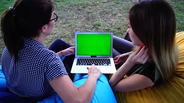 Zwei reizende Freundinnen betrachten Werbung im Internet auf grünem Bildschirm und diskutieren, wie sie an warmen Tagen im Park in weichen Sesseln sitzen. — Stockvideo