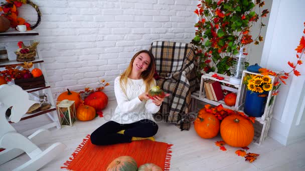 Linda chica posando con calabazas, sonriendo y sentado en el suelo en la habitación brillante en el fondo del paisaje de otoño . — Vídeos de Stock