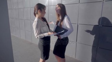İki çekici genç kadın girişimciler tartışmak, belgeleri incelemek ve arka plan açık havada nötr renklerde iş merkezinin üzerinde durmak.