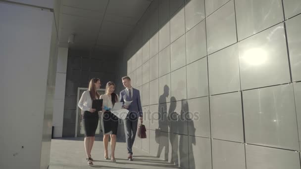 Dos chicas y un chico caminan y conversan con carpetas en las manos en el fondo del centro de negocios al aire libre en colores neutros . — Vídeo de stock