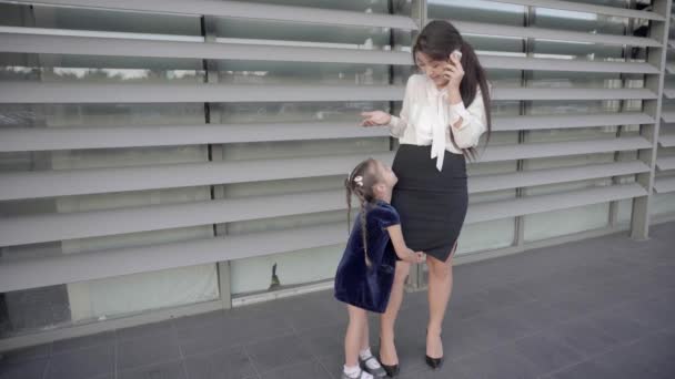 Madre bastante moderna hablando por teléfono, la niña abrazando a la madre y se paran contra la ventana del centro de negocios al aire libre en colores neutros . — Vídeo de stock