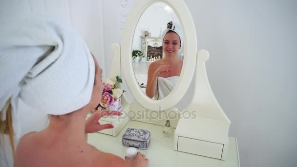 Jong meisje zichzelf bewondert en kijkt naar de reflectie in een spiegel, glimlachen en Open potje crème, zittend op een stoel aan tafel in lichte slaapkamer. — Stockvideo
