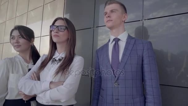 Tres jóvenes empresarios educados sonriendo y posando en la cámara, parados en el fondo de la pared cerca del centro de negocios al aire libre en colores neutros . — Vídeo de stock