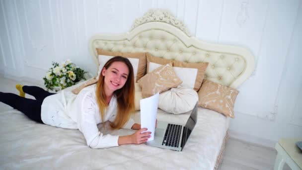 Estudiante mirando la cámara, posando y sonriendo con documentos y papeles en la mano, yace en una cama grande en el dormitorio . — Vídeo de stock