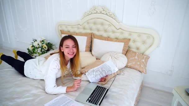 Chica encantadora freelancer utiliza el ordenador portátil para el trabajo, se pone en la cama grande en el dormitorio y sonriendo . — Vídeo de stock