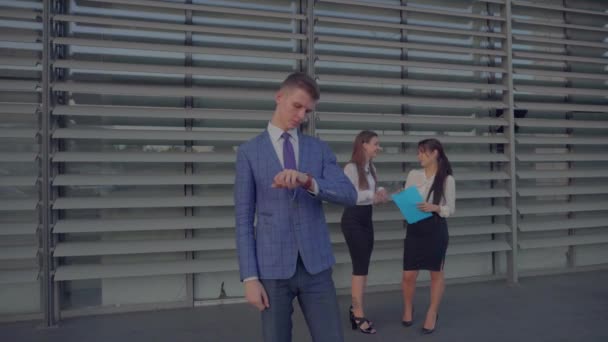 Businessman Stands in Foreground in Front of Camera, Looking at Watch on Background of Colleagues Women Near Business Center in Neutral Colors. — Stok Video