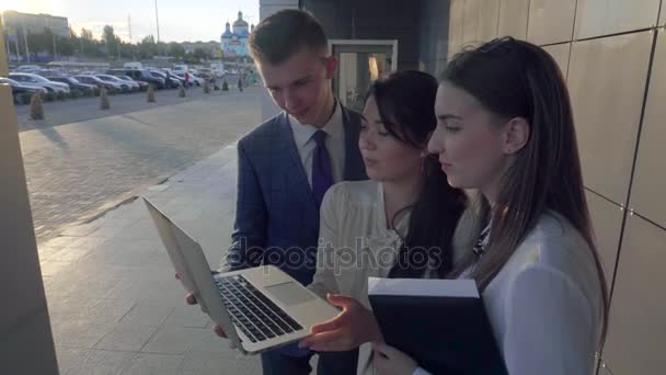Трое молодых студентов, девочки и мальчик работают с ноутбуком и разговаривают на фоне бизнес-центра на открытом воздухе в нейтральных цветах . — стоковое видео