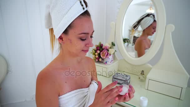 Young Girl Rubs Cosmetic Cream on Face, Looking up and Smiling at Camera in Light Room, Bedroom, Sitting at Table. — Stock Video