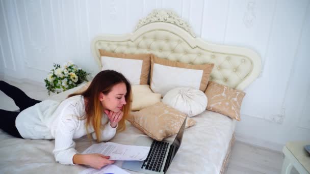 Kobiece uczeń robi pracę domową na komputerze i odczytuje informacje o papiery leżące na duże łóżko w sypialni. — Wideo stockowe