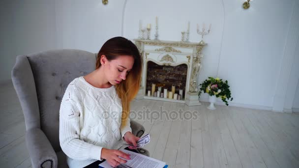 Mujer de negocios moderna lidiando con dinero y lectura Documentación de trabajo, sentada en silla contra la pared blanca y chimenea con velas . — Vídeos de Stock