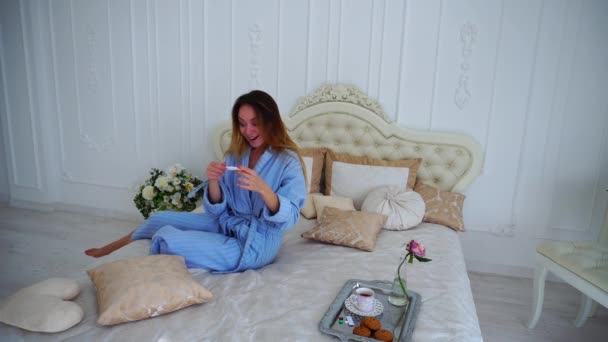 Mujer joven feliz, feliz prueba positiva para el embarazo, sonrisas y muestra la cámara de prueba, sentado en la cama en el dormitorio . — Vídeos de Stock