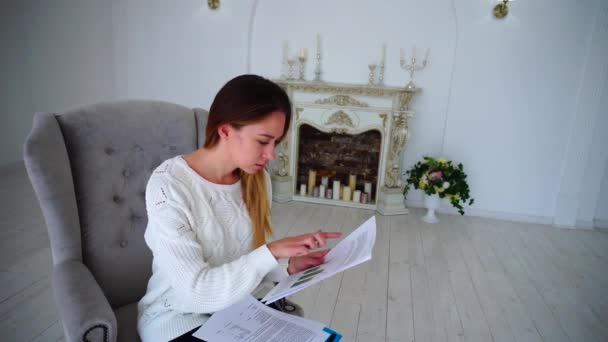 Ragazza aspirante imprenditore analizza con attenzione la carta e gioire del successo, seduto sulla sedia contro il muro bianco e camino con candele . — Video Stock