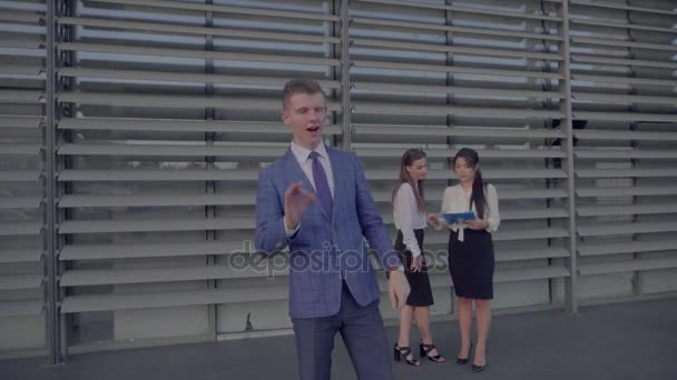 Moderne mannelijk in de voorgrond staat vooraan toont van Camera, gebaren en glimlacht op achtergrond van collega's van Business Center in neutrale kleuren. — Stockvideo