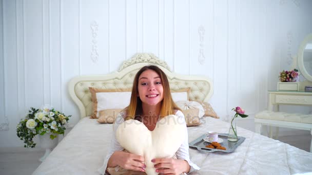 Portrait de femme mignonne tenant oreiller-coeur, posant et souriant tout en s'allongeant sur le lit sur fond de vases avec des fleurs et mur blanc dans la chambre . — Video