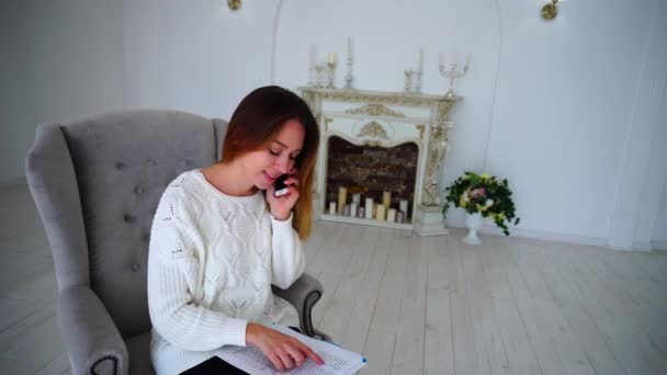 Geschäftige erfolgreiche Geschäftsfrau telefoniert, informiert über Informationen in Zeitungen, sitzt im Stuhl vor weißer Wand und Kamin mit Kerzen. — Stockvideo