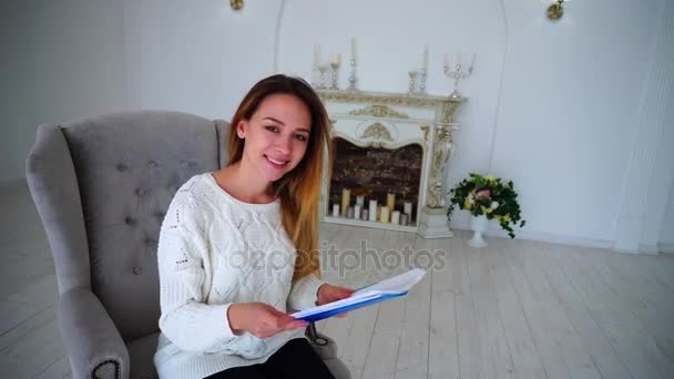 Симпатичная женщина-секретарь держит бумагу и рисование рук, смотрит в камеру и улыбается, сидя в кресле на фоне стены и камина . — стоковое видео