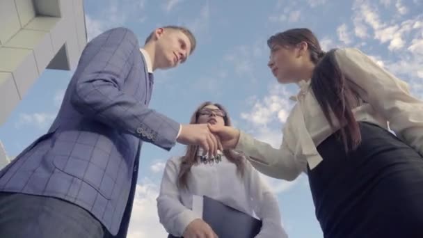Три успішні люди вітають один одного і проведення документи, стоять біля бізнес-центру на тлі неба на відкритому повітрі в нейтральних тонах. — стокове відео