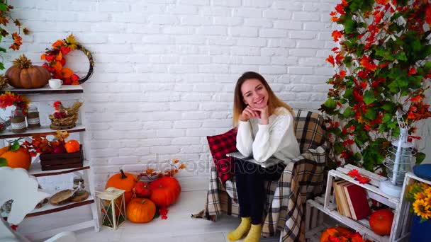 Preciosa chica mirando a la cámara y sonriendo mientras sostiene el gadget en las rodillas y se sienta en la silla en la habitación decorada con decoraciones de otoño . — Vídeos de Stock