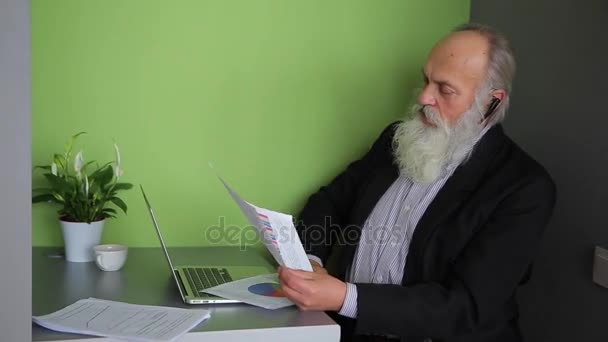 Old Aged Leader Manager Reads Report and Talks on Bluetooth Headset With Boss, Sitting at Table in Computer Cafe During Day. — Stock Video