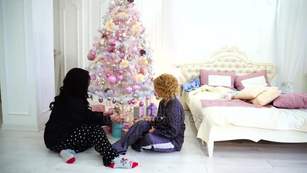 Twee goede vrienden delen aangename emoties en feestelijke cadeaus, zittend op de vloer in lichte slaapkamer in de buurt van bed en kerstboom. — Stockvideo