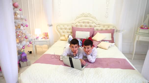 Ragazzi gemelli in posa e giocare sul computer portatile, sdraiati sul letto in camera luminosa con albero di Natale di giorno . — Video Stock