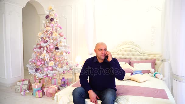 Mann gratuliert Verwandten telefonisch mit Neujahrsurlaub mit Söhnen, sitzt am Wintertag auf Bett im Schlafzimmer mit festlichem Weihnachtsbaum. — Stockvideo